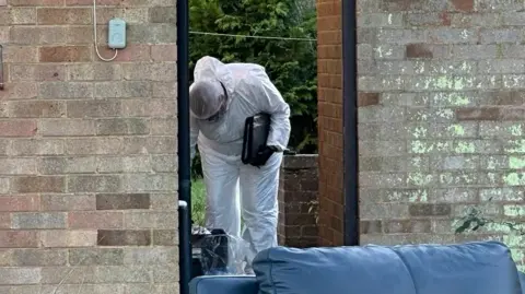 Ben Parker/BBC A forensic officer dressed in a white suit with a white hair net is pictured leaning over looking at something toward the ground in the side garden of a home which is behind a brick wall with an open fence door.