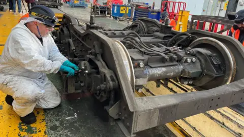 An engineer in white overalls is crouched down working on a wheel with a spanner.