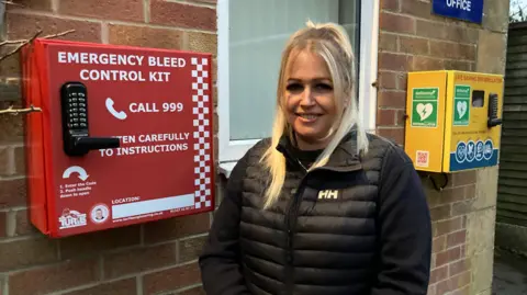 Anya stands outside Cadbury Hall, with the bleed kit in a large red box affixed to the brick wall. The cabinet says "emergency bleed control kit, call 999", with a yellow defibrillator cabinet behind her.  She has long blonde hair, tied back in a ponytail, and wears a black puffer coat. 