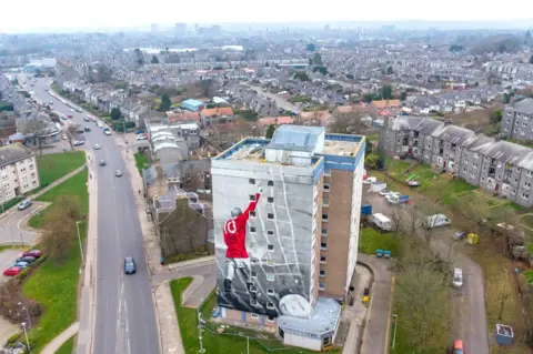 Michael Traill Painted image of the late footballer Denis Law in a red shirt, number 10 on back, arm raised, on the side of a high rise building, with Aberdeen in the background.