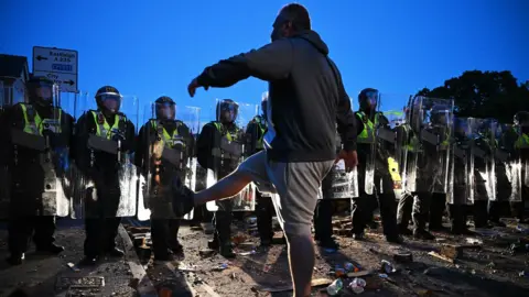 Police in Southampton line up in a clash against protesters . A male protester is seen in the foreground silhouetted against the sky kicking his leg at the police 