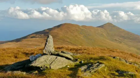 Rolling green hills, with some orange coloured patched. Some large stones can been seen in the foreground. The sky is blue and has white fluffy clouds.