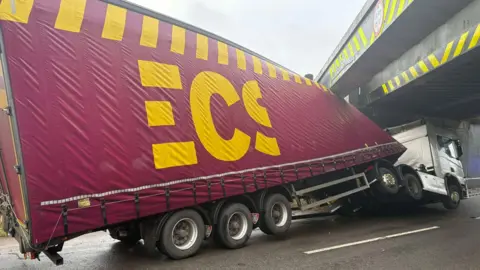 An image of a large lorry that has been involved in an accident underneath a bridge. The truck, which is maroon and yellow, is angled and appears to have been damaged on impact and displaced.