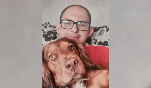 Jack Nixon, in a red t-shirt and black-rimmed glasses, is sitting on a couch and smiling at the camera while cuddling a dog. 