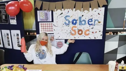 Charlie Fae Charlie in university - she has long blonde hair and is wearing a white jumper with a blue logo on it. She is wearing an orange face mask and is holding a peach coloured water bottle with her arms in the air. She is gesturing to a blue board which has a white piece of paper on it that reads "sober soc" in blue and orange writing.
