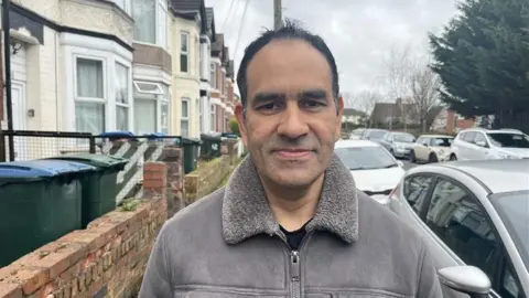 A man with dark hair is standing in a street with a row of houses and a row of parked cars behind him. He is wearing a grey jacket with a fluffy collar. There are trees and further houses in the distance.