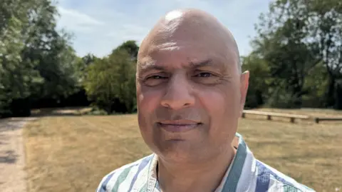 BBC Harjit Singh Nijjer is smiling. He is standing in the park with trees behind him.