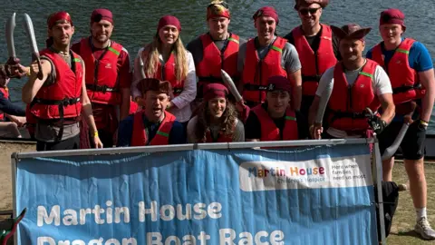 BBC/Jonathan Reed A group of people standing together by a body of water, posing behind a large blue banner that reads “Martin House Dragon Boat Race”. The group is wearing red life jackets and headscarves, and several people are holding paddles.
