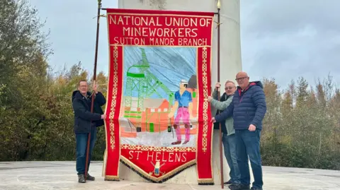 Three men hold a traditional union banner between them. It is twice their height, which is red around the edges and says National Union of Mineworkers Sutton Manor branch at the top and St Helens at the bottom. It shows a miner with helmet and head lamp in front of a factory and mill apparatus.