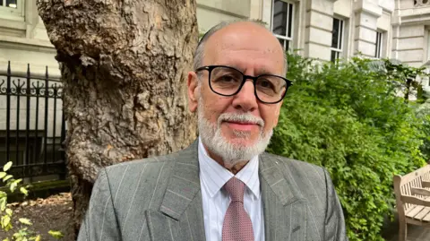 George Psaradakis in a mauve tie, grey suit and black glasses standing in a park with a tree trunk behind him.