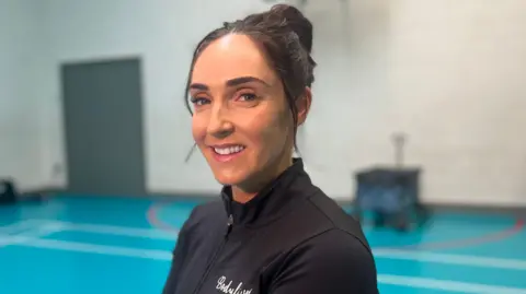 BBC Aimee is smiling at the camera. She has dark brown hair and is wearing a black sports jacket. She is sitting down on a chair in the middle of an indoor sports court. 