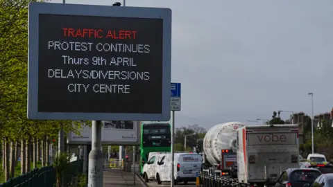 PA Media On the left, a sign which reads: 'TRAFFIC ALERT - PROTEST CONTINUES - Thurs 9th APRIL - DELAYS / DIVERSIONS - CITY CENTRE'. Behind the sign there is a road which appears to have a traffic jam. 