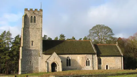 Evelyn Simak/Geograph All Saints' Church, West Harling. Its flint-built tower is on the left and the body f the church runs to the right. There is a porch in front of its entrance, to the right of the tower. There are trees behind it and blue and cloudy sky above.