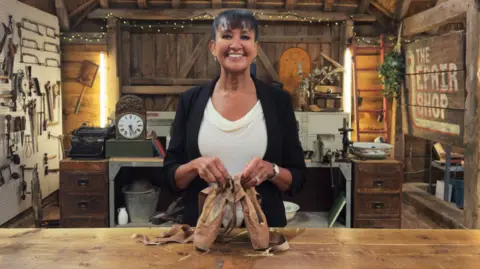 A woman smiling into the camera holding ballerina shoes. She is smiling and has a fringe. She is wearing a white top and has a black blazer on. She is in a workshop where they have different antique items on display. 