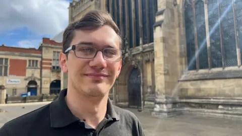 Amanda White/BBC A man standing in front of Hull Minster. He has dark wavy hair and is wearing a black shirt and glasses.
