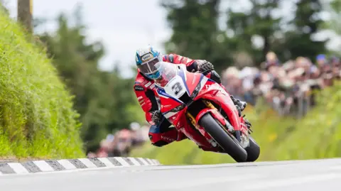 ISLE OF MAN TT Dean Harrison on a red superbike with the number 3 on the front. He is rounding a bend that had a black and white kerb and there are spectators watching in the background.