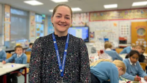 Sarah smiles at the camera as she wears a dark dress with floral patterns on it. She has a blue school lanyard around her neck. Her dark blonde hair is tied into a bun behind her head. Behind her children are working on making hampers. 