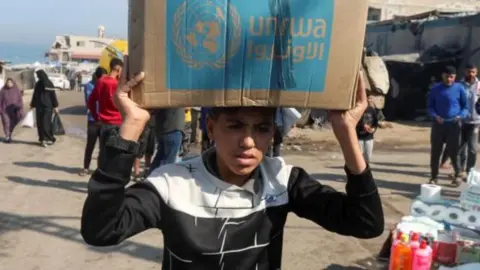 A teenage boy carries a cardboard box with the words "Unrwa" on them