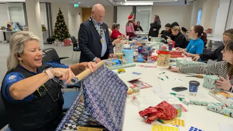 Epping Forest District Council A woman in police uniform wrapping a present on a long, white table with several volunteers in the background doing the same.