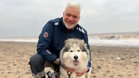 Adrian Ashworth is wearing a navy jacket and dark grey trousers. He has white hair, a grey beard, and tattoos on his hands. He is smiling brightly, knelt on a beach and cuddling a large grey/white husky dog who has dark eyes, and his mouth wide open.