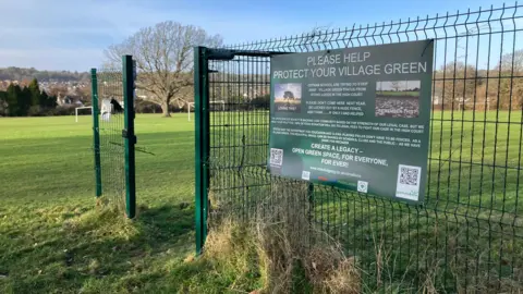 A green fence is seen along a green field. There are goal posts in the distance. A sign on the fence says Please help protect your village green.
