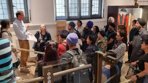 A little History of the Sikhs Tour leader Stephen Barker talking in front of attendants inside a building. Some colourful clothes can be seen in a room behind the.