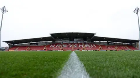 A pitch level view of the Stok Cae Ras ground, with a grandstand and two floodlights in the distance. 