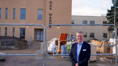 Luca Felice/Milton Keynes University Hospital NHS Foundation Trust A man in a suit stands in front of metal fencing at a hospital construction site, with the words OAK WARDS visible on the building behind them. Construction materials and equipment are scattered around the fenced-off area.