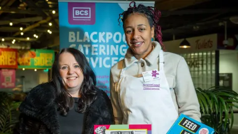 Blackpool Council Two women smile for the camera in front of a sign saying Blackpool Catering Services. On the right, the taller woman is wearing a white apron over her stone coloured jacket. The shorter woman on the left has black hair and a black jacket