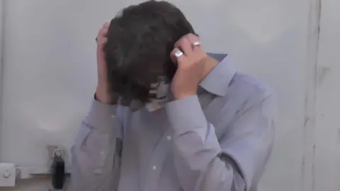 Pollyfromthedirt Pollyfromthedirt is wearing a blue shirt and he has lowered his head with his hands on both sides of his face. He is wearing two silver rings on his right hand.