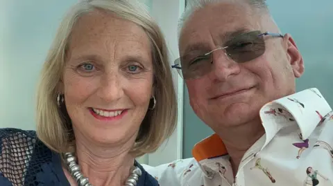 Gordon Hollingsworth A woman and a man appear together in the photo. The man has short grey hair and he is wearing shaded glasses. The woman has blonde hair and is smiling