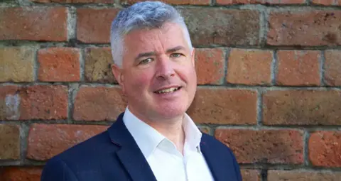 Scottish Information Commissioner A man with grey hair, wearing a blue suit jacket and open-necked shirt, stands in front of a brick wall