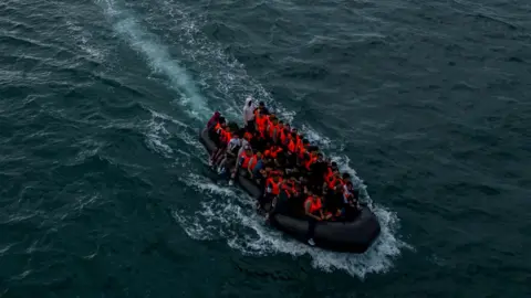 An inflatable dinghy full of people wearing lifejackets makes its way through water 