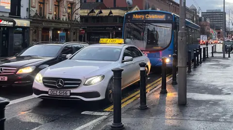 A while Mercedes Value Cabs taxi driving in a bus lane on Belfast's Great Victoria Street before Christmas. A bus with a sign that says "Merry Christmas" is behind the taxi.