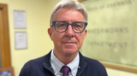 Deputy Mark Helyar - A man with grey hair, wearing blue spectacles. He has a blue jacket on with a blue and white checked shirt. 