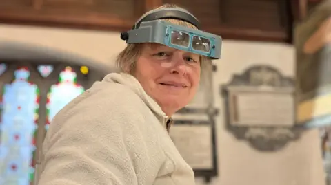 A woman with grey hair and wearing a magnifying head set is half-smiling at the camera. She is wearing a warm fluffy jumper and you can see the stained-glass windows and plaques in the church
