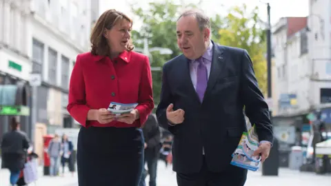 Getty Images Two people in the centre of the photograph are talking and holding leaflets. To the left is Leanne Wood, who is wearing a dark skirt and a red jacket, and to the right is Alex Salmond, who is wearing a suit, a pink short and purple tie. 