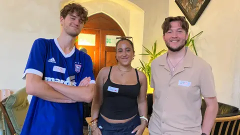 Jon Wright/BBC Three young people stand in a row, inside a church with sofas in the background. Jack has a blue Ipswich football jersey on, Freya wears a black strap top and Adam wears a beige polo shirt with a flopping brown middle-parted fringe.