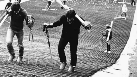 Kendal Snowsports Club A black-and-white photo of skiis on a dry slope - dated in the 1980s. There are two men walking up a slop wearing boots and have their skiis resting on their shoulders and have a pair of poles in one hand. The dry slope matting is very basic. 