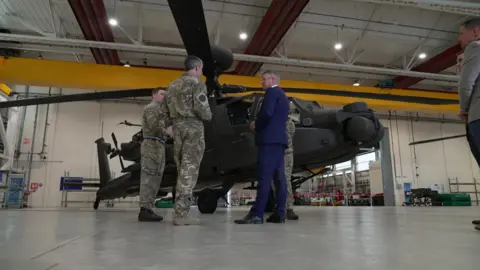 Luke Pollard MP wearing a blue suit stood talking to armed forces personnel in front of an Apache helicopter.