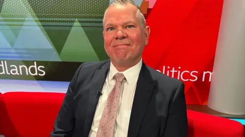 A man wearing a suit and pink tie sits on red sofa looking at the camera. A screen can be seen behind him.