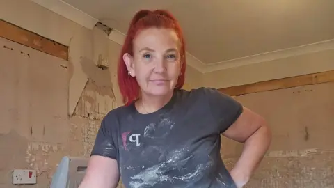 Steph Leese, who is wearing a grey top covered in plaster, is standing in a room which needs plastering. 