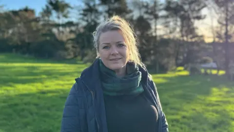 Kirree, a woman with a blonde ponytail, she smiles and wears a scarf and navy puffer jacket as she stands in a field.