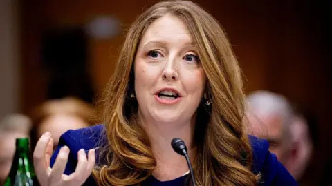 Close-up of Casey Means, wearing a blue shirt with long reddish hair, speaking into a microphone