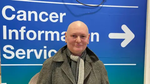 The image shows a bald man in a grey tweed-style overcoat and a dark-and-light grey scar around his neck, standing in front of a sign saying 'Cancer Information Services'.