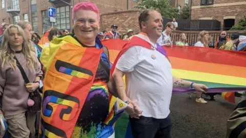 KIT TAYLOR/BBC People taking part in the parade, wearing outfits filled with different colours and patterns. One person has pink hair, holding hands with another person next to them.