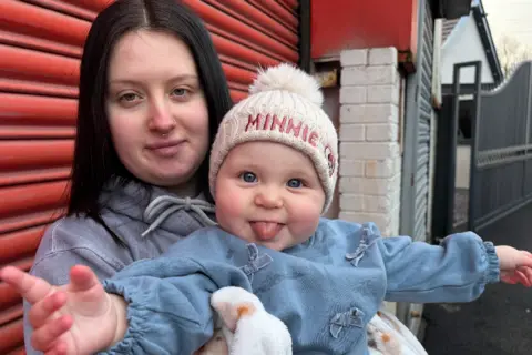 Close up of Alex holding Lilah in front of a red garage. Lilah has a big smile and is looking at the camera with her tongue out. 