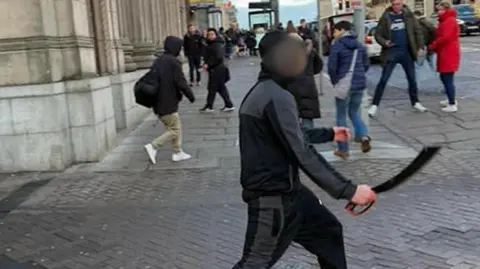 Person holding a large bladed weapon walking across a city centre street, with pedestrians nearby and buildings lining the pavement.