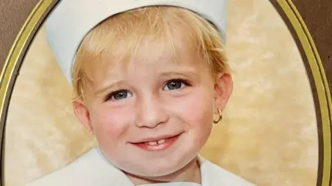 Nunan family James Nunan pictured as a toddler. He has blond hair and is wearing a white sailor's hat. He has a gold earring in his left ear and is smiling.