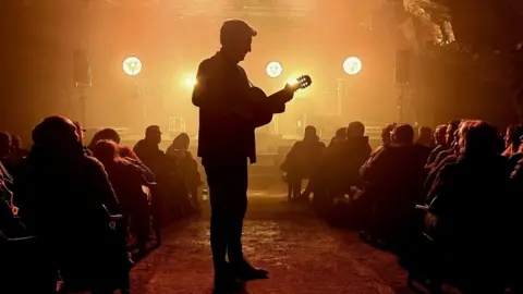 Carnglaze Caverns A silhouette picture of a man playing a guitar in a dark underground room. There is a stage with some lights in the background as well as a crowd of people all sat down. 